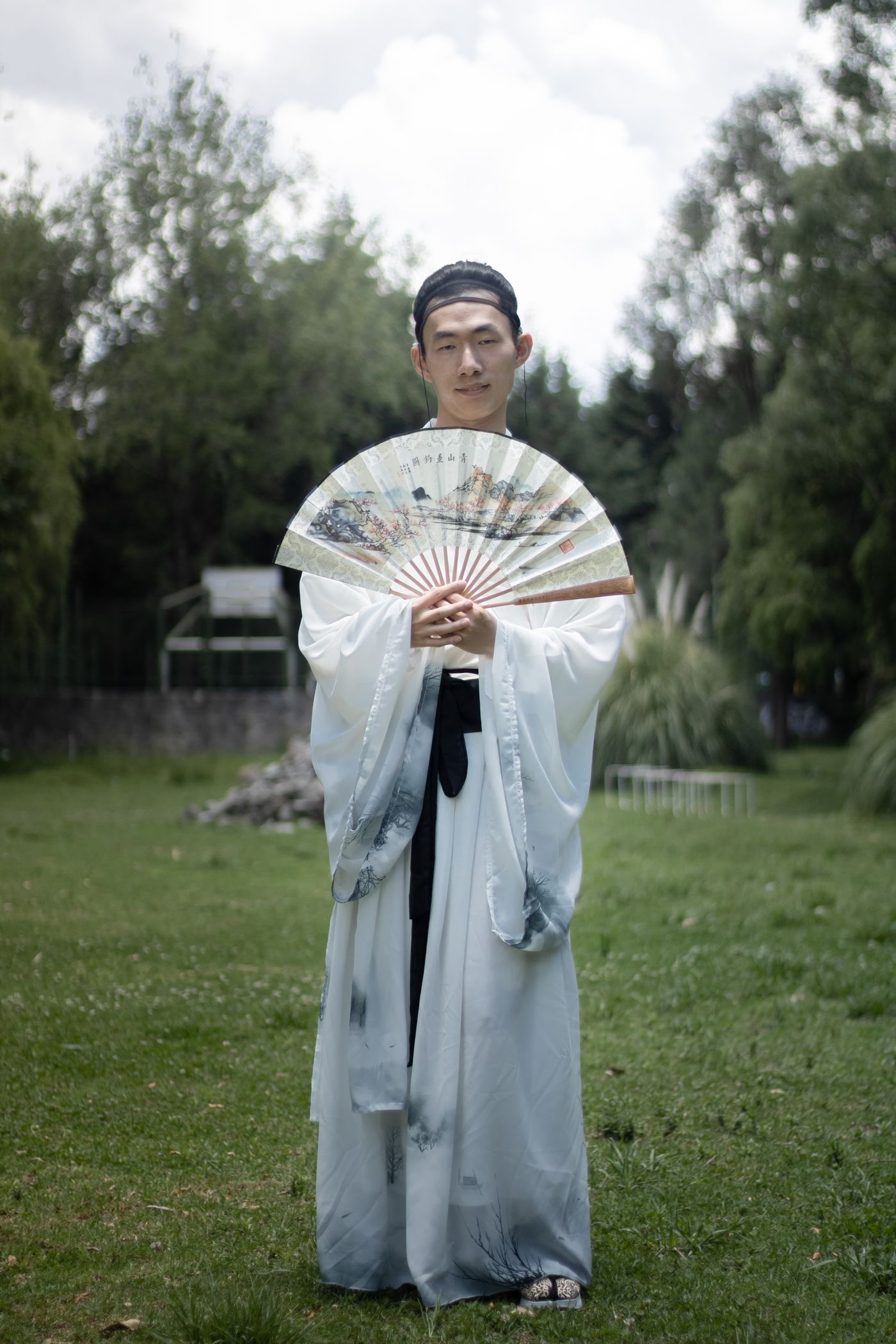 Man in traditional Hanfu holding a folding fan in a peaceful park