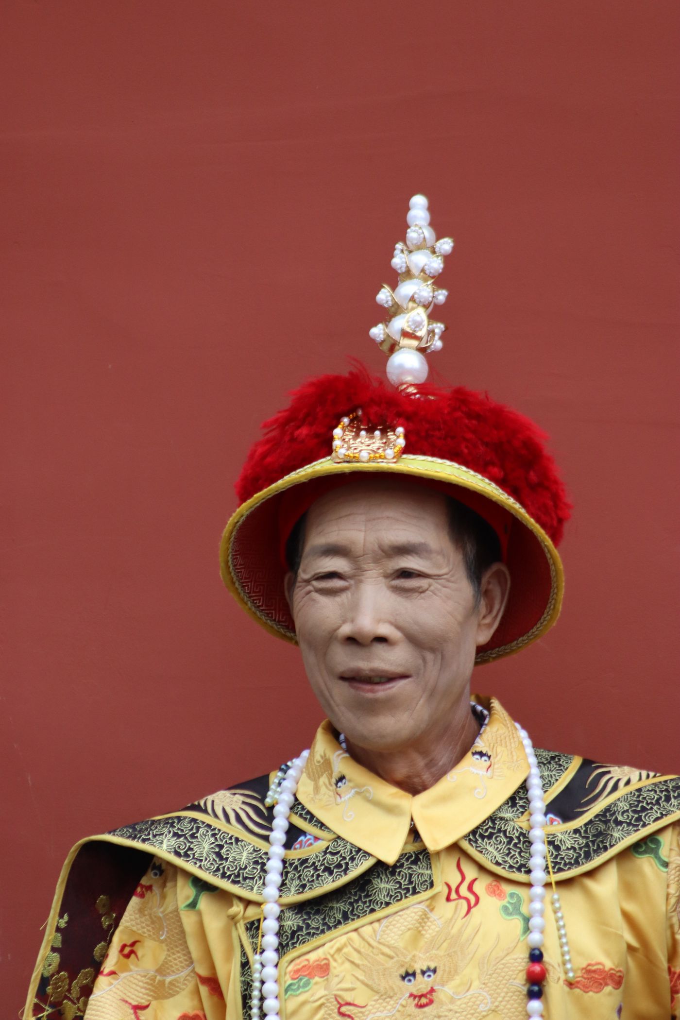 Man wearing traditional imperial-style Chinese robe with elaborate headdress against a red backdrop